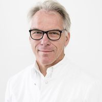 Porträt eines älteren Mannes mit grauem Haar, schwarzer Brille und weißem Hemd vor hellem Hintergrund
