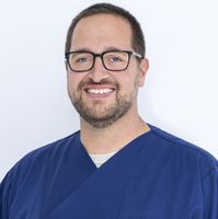 Lächelnder Mann mit Brille und Bart in dunkelblauer medizinischer Kleidung vor weißem Hintergrund