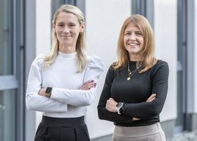 Drei lächelnde Frauen in Businesskleidung stehen mit verschränkten Armen vor hellem Hintergrund.